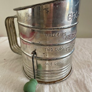 May include: A vintage silver-colored metal flour sifter with a green handle. The sifter has markings for one, two, and three cups of flour.