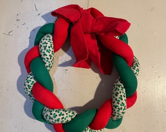 May include: A red and green Christmas wreath made of fabric with a red bow. The wreath is braided and has a pattern of green holly leaves on a white background.