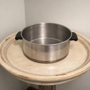 May include: A round, stainless steel cooking pot with a polished metallic finish and two black, heat-resistant side handles. The pot features a wide, open top and a flat base, designed for stovetop use in a kitchen setting for various cooking tasks.