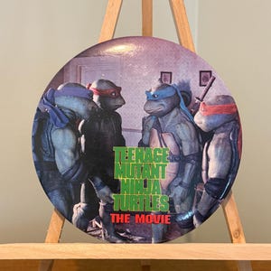 May include: A round Teenage Mutant Ninja Turtles movie button featuring the four turtles in their signature colors. The button has the movie title in green and red text.