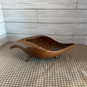 May include: A handcrafted wooden bowl with an unusual, organic shape. The bowl is a warm brown colour and rests on three small legs. The interior is carved, and the overall design suggests a natural, flowing form. The bowl is set on a brown and white checkered surface.
