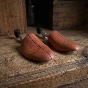 May include: A pair of reddish-brown wooden shoe trees featuring a smooth, contoured toe section and a metal spring mechanism with a black plastic heel knob, designed to maintain the shape of footwear while providing structural support.
