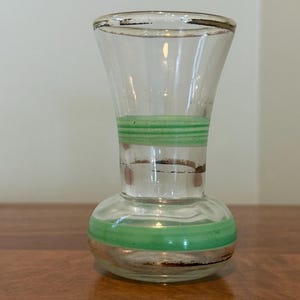 May include: A clear glass vase with a flared rim and a rounded base, featuring two horizontal bands of light green paint around the neck and body, along with thin, worn metallic gold trim accents on the rim and near the painted sections.