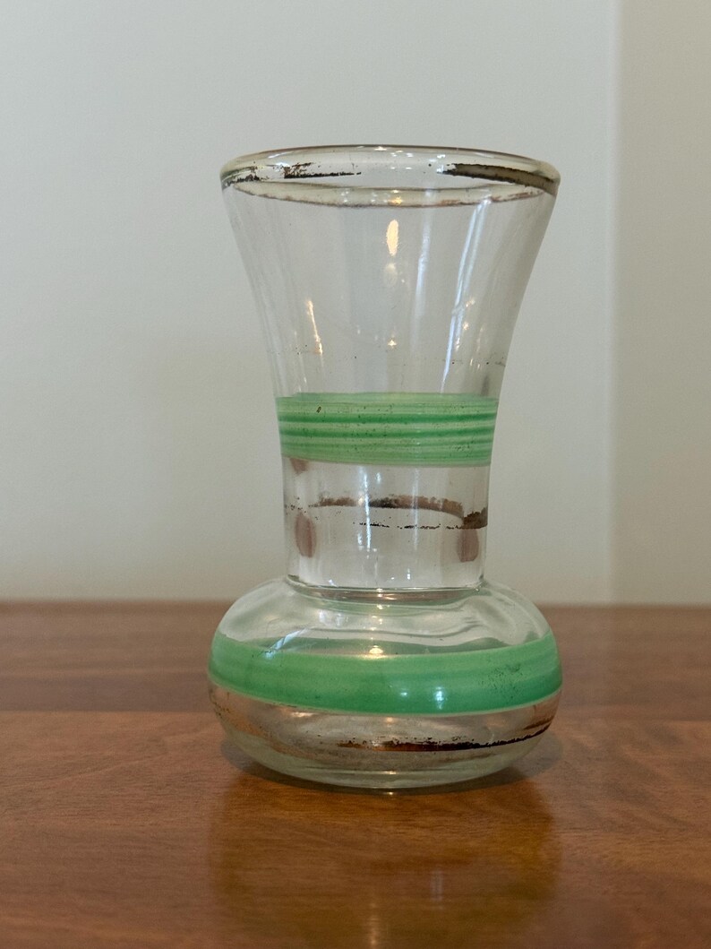 May include: A clear glass vase with a flared rim and a rounded base, featuring two horizontal bands of light green paint around the neck and body, along with thin, worn metallic gold trim accents on the rim and near the painted sections.