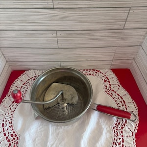 Peut inclure: Un moulin à légumes vintage en acier inoxydable avec une poignée et un bouton rouges. Le moulin repose sur un napperon en dentelle blanche sur une surface rouge. Le moulin à légumes est conçu pour réduire en purée les fruits et légumes.