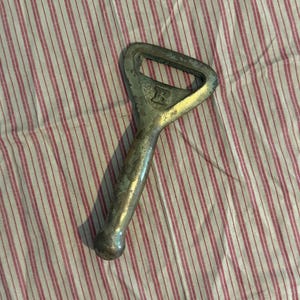 May include: A vintage metal bottle opener with a handle and a loop for leverage. The opener has a worn, silver finish and is resting on a red and white striped fabric. The opener has the letter "R" on it.