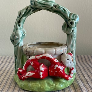 May include: A ceramic planter shaped like a well with a green vine handle and a red elf figure lying on the ground.