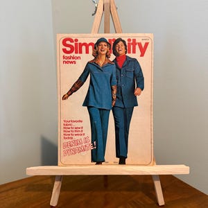 May include: A vintage magazine cover titled "Simplicity fashion news" featuring two people in denim outfits. The cover has the text "DENIM IS DYNAMITE!" and other text about sewing. The magazine is displayed on a small wooden easel.