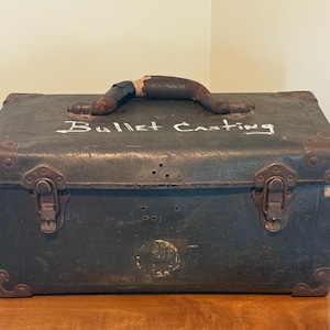 May include: An antique, dark green metal box with the words "Bullet Casting" in white script. The box has a worn, brown handle and metal corner protectors. Two metal latches secure the front.