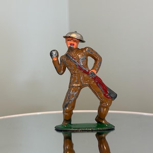 May include: A vintage toy soldier figurine in a brown uniform, holding a grenade and a rifle. The soldier wears a silver helmet and has orange facial features. The figurine stands on a green base, with visible wear and tear.