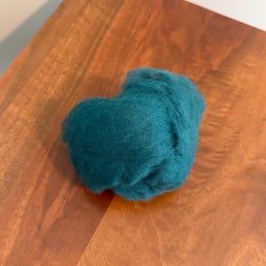 May include: A teal ball of wool fibre rests on a wooden surface. The wool has a soft, fluffy appearance and an irregular shape. The wood grain is visible, adding a natural texture. The scene has a simple, tactile aesthetic, with a focus on the wool's texture.