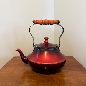 May include: A vintage red and black tea kettle with a wooden handle and a black metal handle. The kettle is sitting on a wooden surface. The kettle has a spout and a lid.