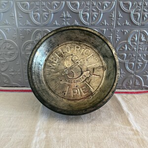May include: A vintage, round metal pie tin with the text "MELLO RICH" and "MRS SMITH'S PIE" embossed on the surface. The tin has a distressed, aged appearance with a silver and dark gray color scheme. The background features a patterned, silver-colored wall.