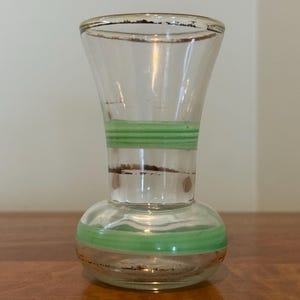 May include: A clear glass vase with a flared top and a rounded base, featuring two horizontal light green bands encircling the body. The rim and the middle section of the vase are accented with thin, worn brown metallic trim details.
