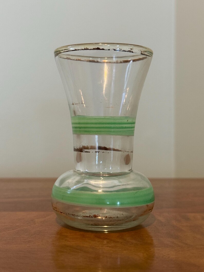 May include: A clear glass vase with a flared top and a rounded base, featuring two horizontal light green bands encircling the body. The rim and the middle section of the vase are accented with thin, worn brown metallic trim details.