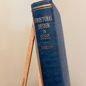 May include: A blue book titled "STRUCTURAL DESIGN IN STEEL" by Shedd. The book is propped up on a wooden stand. The title is in gold lettering.