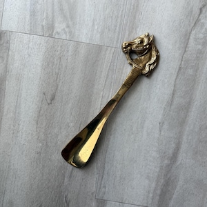 May include: A brass shoe horn with a horse head design. The horse head is at the top of the handle, with a detailed mane and bridle. The horn has a curved, elongated shape.