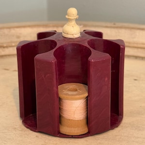 May include: A burgundy plastic spool holder with six slots for spools of thread. A white plastic spool of thread is in one of the slots. A white plastic chess piece sits on top of the holder.