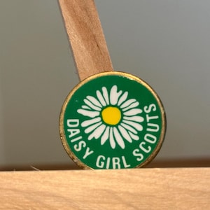 May include: A circular metal pin with a green background featuring a white daisy flower with a yellow center. The text DAISY GIRL SCOUTS is printed in white capital letters around the bottom half of the flower design on the green enamel surface.