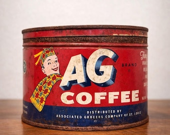 May include: A vintage red and blue cylindrical metal coffee tin featuring the text AG COFFEE and a graphic of a person wearing a yellow and red plaid hat and scarf. The container shows signs of rust along the top rim and base edges.