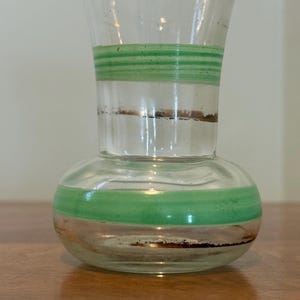 May include: A clear glass vase with a bulbous base and a flared neck, featuring horizontal green bands and thin metallic copper-coloured rings encircling both the lower body and the mid-section of the glass vessel.