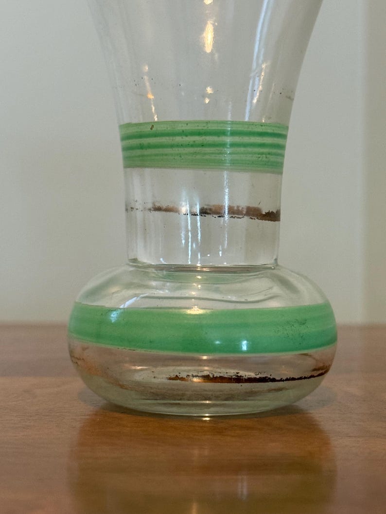 May include: A clear glass vase with a bulbous base and a flared neck, featuring horizontal green bands and thin metallic copper-coloured rings encircling both the lower body and the mid-section of the glass vessel.