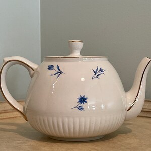 May include: A white teapot with a gold trim and blue floral designs. The teapot has a round body and a spout on the right side.