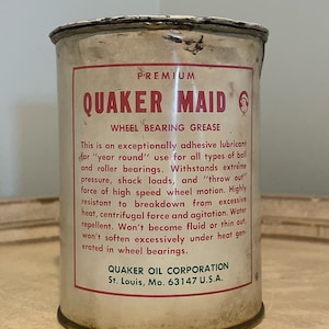 May include: A white metal can with a red and white label. The label reads "PREMIUM QUAKER MAID WHEEL BEARING GREASE". The text on the label describes the product as an exceptionally adhesive lubricant for "year round" use for all types of ball and roller bearings. The label also states that the product is water repellent and won't become fluid or thin out under heat generated in wheel bearings. The label also includes the company name and address: "QUAKER OIL CORPORATION St. Louis, Mo. 63147 U.S.A."
