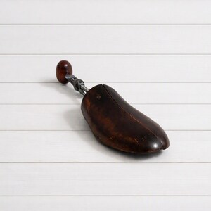 May include: A dark brown wooden shoe tree with a smooth, polished finish. The device features a rounded heel knob attached to a silver-toned metal spring mechanism that connects to the main foot-shaped wooden form for maintaining shoe shape.