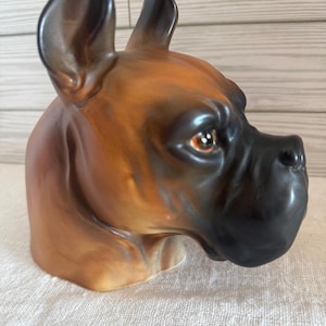May include: A ceramic Boxer dog head sculpture. The sculpture features a glossy finish with brown, black, and tan coloring. The dog's ears are erect, and its eyes are open. The sculpture is set on a white surface.