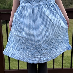 May include: A blue and white gingham apron with black embroidered details. The apron has a pocket on the front and a gathered skirt.