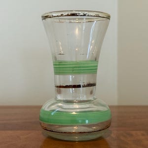 May include: A clear glass vase with a flared rim and a rounded base. The vase features horizontal bands of light green paint and thin, dark brown metallic-toned stripes around the neck and the lower body, with visible wear on the rim.