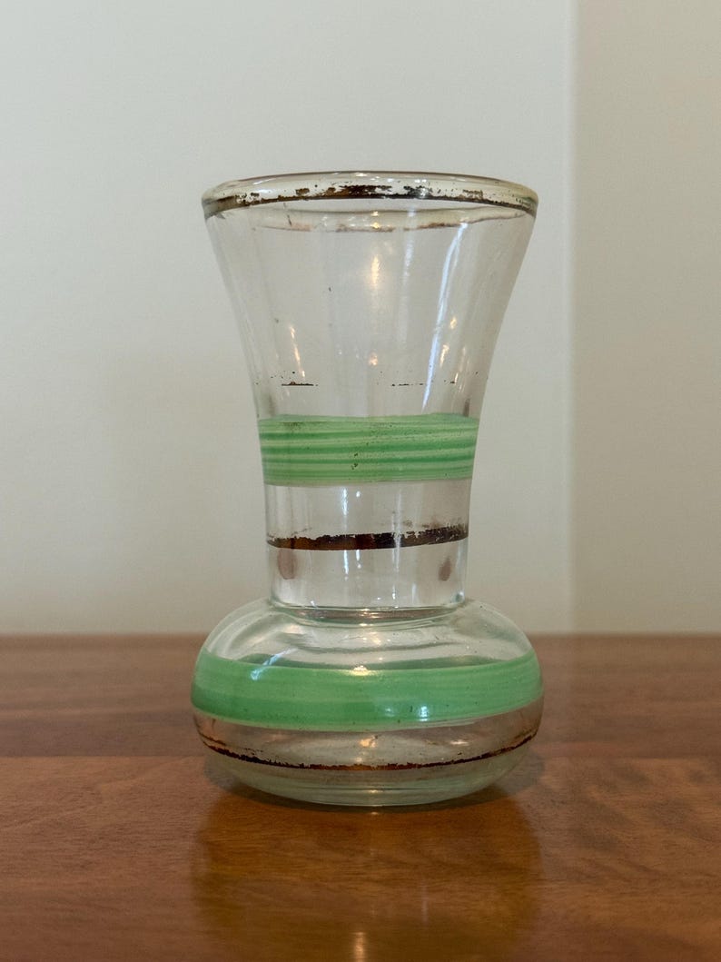 May include: A clear glass vase with a flared rim and a rounded base. The vase features horizontal bands of light green paint and thin, dark brown metallic-toned stripes around the neck and the lower body, with visible wear on the rim.