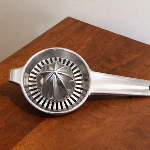 May include: A silver-colored metal citrus juicer featuring a central reamer cone surrounded by a circular strainer with vertical slots. The tool includes a long, flat handle with a small circular hole at the end for hanging storage.