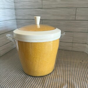 May include: A vintage ice bucket with a yellow woven texture. The bucket has a clear lid and a white rim and handle. The lid has a white handle. The bucket is sitting on a striped fabric.