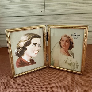May include: Two vintage gold-framed photo holders. Each frame holds a portrait. The right frame has text that reads "#7D 3 1/4 x 4 1/4 MADE IN U.S.A.". The frames are hinged together.