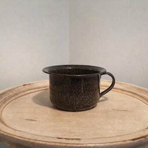 May include: A dark brown, speckled enamel mug with a wide, flared rim and a single curved handle. The mug features a mottled, textured finish throughout its exterior surface, providing a rustic appearance. The design includes a deep, cylindrical body.