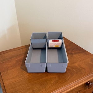 May include: A light gray plastic drawer organizer set featuring two long rectangular bins and two smaller square bins that sit inside. A white and red Rubbermaid label is attached to one of the square bins, displaying the text Instant Drawer Organizer.