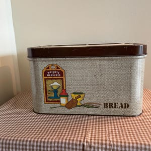 May include: A vintage brown and beige metal bread box with a burlap-like texture. The front features a graphic of a scale, a jar, a wooden rolling pin, and a bowl. The text "Weights Measures" and "Bread" are printed on the box.