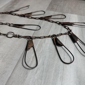 May include: Close-up of several antique metal chains with large, open wire loops and clasps. The chains and loops have a weathered, rusty appearance, suggesting age and use. The background is a light gray wooden surface.