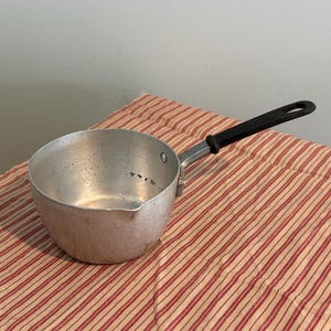 May include: A small, aluminum saucepan with a black handle. The pan has a pouring spout and is sitting on a red and white striped cloth. The pan is likely used for cooking or heating liquids.