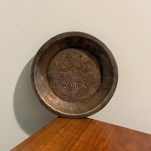 May include: A small, round, metal pie pan with a textured starburst pattern in the center. The pan has a dark, aged appearance, with the text "OVENE N306-6 USA" embossed on the bottom.