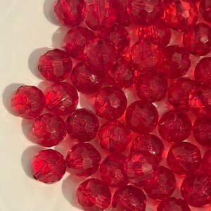 May include: A collection of faceted, round, red beads. The beads are scattered on a white surface. Each bead has multiple facets that catch the light, creating a sparkling effect. The beads are a vibrant, transparent red color.
