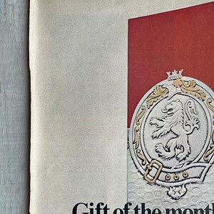May include: A vintage poster advertising a gift of the month club. The poster features a red background with a silver crest featuring a lion and the text "Gift of the month".