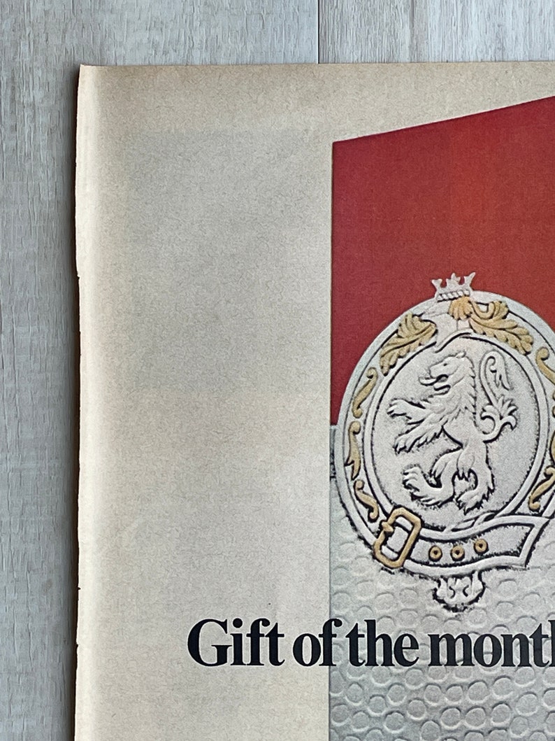 May include: A vintage poster advertising a gift of the month club. The poster features a red background with a silver crest featuring a lion and the text "Gift of the month".