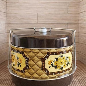 May include: A round cake carrier with a dark brown lid and base, featuring a central band of quilted gold fabric decorated with a yellow and white floral pattern. The container includes a silver metal handle and a white knob on the lid.