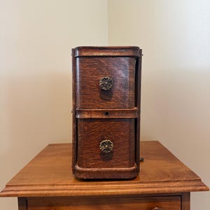 May include: A vertical wooden storage unit featuring two stacked drawers with a dark brown wood grain finish. Each drawer is equipped with a round, metallic floral-patterned knob. The unit has rounded corners and a compact, rectangular design.
