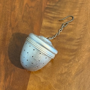 May include: A silver tea infuser with a perforated, egg-shaped body and a hinged lid. A small chain and hook are attached to the top. The infuser is designed to hold loose tea leaves for steeping.