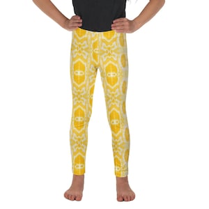 Pode incluir: Uma menina com uma camiseta preta e leggings amarelas. As leggings apresentam um padrão repetitivo branco e amarelo. A menina está em pé com as mãos nos quadris, sorrindo para a câmera. O fundo é branco.