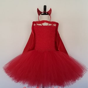 May include: A red devil costume set displayed on a mannequin. The set includes a tutu dress, cape, and a headband with glittery horns. The dress has a crochet bodice and a full, layered tulle skirt. The cape is a solid red fabric.